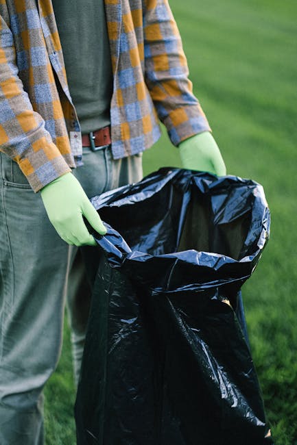 A person wearing a plaid shirt in shades of yellow, gray, and blue, along with green rubber gloves, is holding open a large black rubbish bag made of shiny plastic material. The bag is partially unfolded, positioned in front of the person with the open top revealing its empty interior. The individual is standing outdoors on a grassy area with a blurred green background, likely a lawn or garden, and their waist and legs are visible, dressed in light-colored trousers and a brown belt. The scene suggests an activity related to waste collection or rubbish disposal, consistent with private or independent rubbish removal services often employed to avoid hidden fees. The natural lighting highlights the textures of the bag’s glossy surface and the fabric of the clothing, emphasizing the practical aspect of rubbish handling in an outdoor setting, which could be part of an on-site clearance or waste management process.
