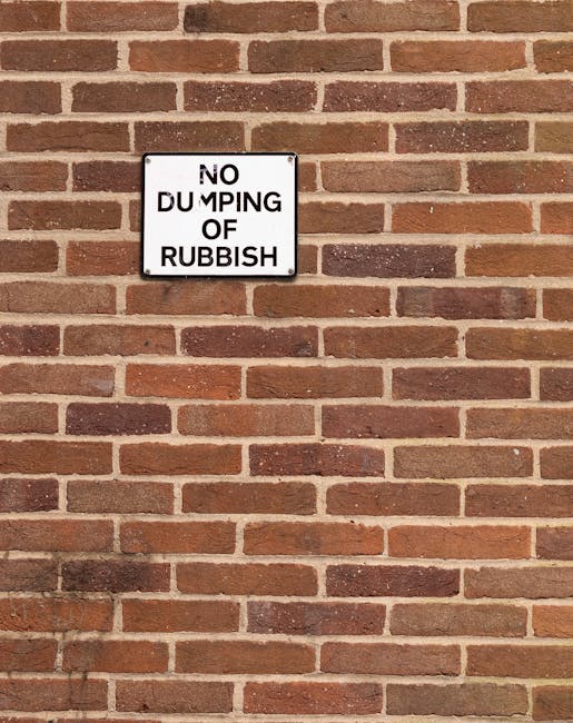 A rectangular metal sign mounted on a red brick wall displaying the message 'NO DUMPING OF RUBBISH' in black capital letters on a white background. The bricks are arranged in a horizontal pattern with varying shades of reddish-brown and light brown, and the mortar lines are clearly visible. The sign is positioned towards the upper left side of the image, with the wall extending uniformly in all directions, highlighting a typical external urban environment where waste management regulations are enforced to prevent illegal rubbish disposal, supporting the context of private rubbish removal services like those offered by rubbishremovalwesthampstead.co.uk.