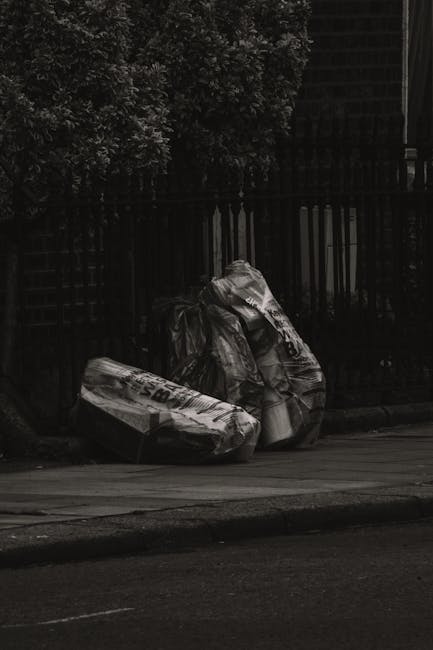 Three large black plastic rubbish bags filled with waste material are placed on the pavement beside a black metal fence, which encloses a garden area with dense foliage and branches at the top. The bags appear crumpled and are positioned close to each other, with one slightly leaning against the fence. The scene is set outdoors on a street with an asphalt road in the foreground and a concrete curb separating the pavement from the road. The lighting suggests a shaded area, with the background consisting of a dark, shadowed fence and leafy greenery. The overall setting reflects a typical private waste disposal scene suitable for local rubbish removal services or independent collection methods, exemplifying alternatives to council-led bulky waste collection, as might be managed by companies specializing in rubbish removal in West Hampstead.