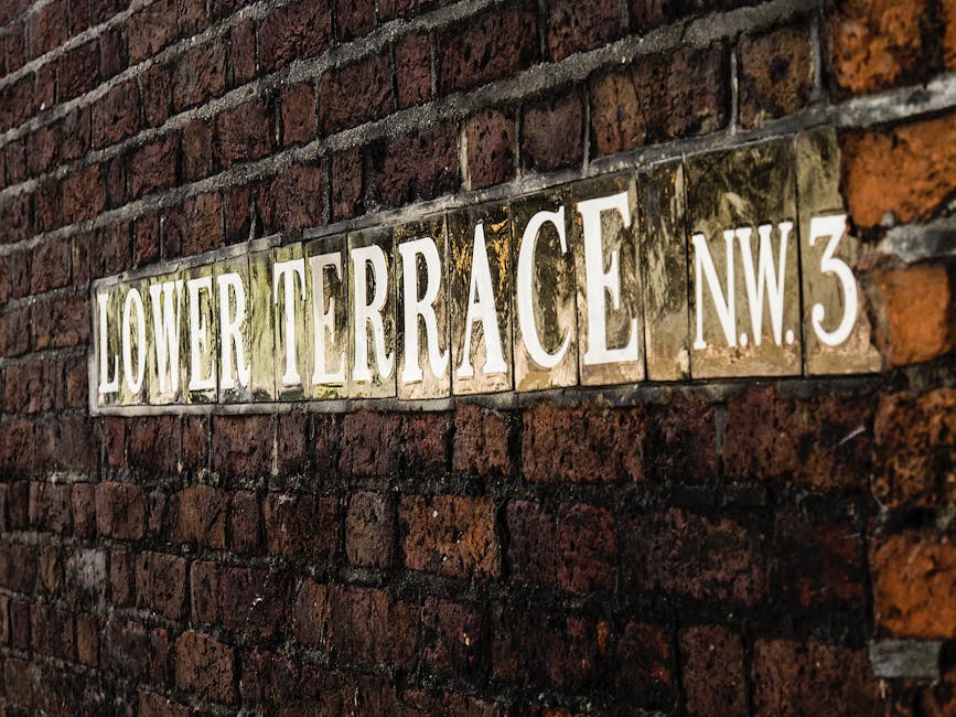 A close-up of a weathered brick wall with a vintage street sign mounted on it, indicating the intersection of Branch Hill and West Heath Road. The sign is composed of black tiles with white embossed lettering, some of which show slight wear and moss growth, reflecting age and outdoor exposure. To the right of the sign, there is a small white hand pointing to the right, also affixed to the wall. The background features moss and lichen growth on the bricks, suggesting an environment that experiences regular humidity. The wall's bricks are dark, uneven in tone, and vary in size, with some bricks appearing more glazed or textured than others, contributing to a rustic and historical appearance. The lighting is natural, emphasizing the textures of the moss and the aged surface of the sign, which provides a neutral, professional look typical of outdoor street signage. The scene visually relates to urban context where private property or local authority signage might complement or indicate access points where rubbish removal or waste management services are needed for surrounding residential or historical areas.
