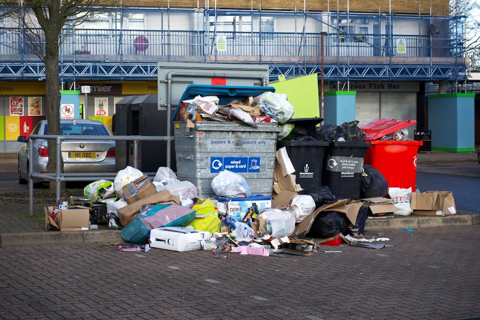 A large accumulation of mixed household waste, including cardboard boxes, paper, plastic bags, and black recycling sacks, is piled around a grey communal rubbish skip on a paved urban street. The skip, partially open, is filled with overflowing paper and cardboard, with some materials spilling onto the surrounding area. To the left of the skip, a small silver car is parked behind a metal railing, and a leafless tree with a slender trunk is visible in front. In the background, there is a building with storefronts, painted in yellow, with signage indicating retail shops, and a blue scaffolding structure covering the upper level of the building. The scene is illuminated by natural daylight, and the overall environment suggests an area experiencing delays in rubbish collection or a private waste disposal point, managed or serviced by [COMPANY_NAME], with the focus on unconventional waste handling outside standard municipal collection times or procedures.