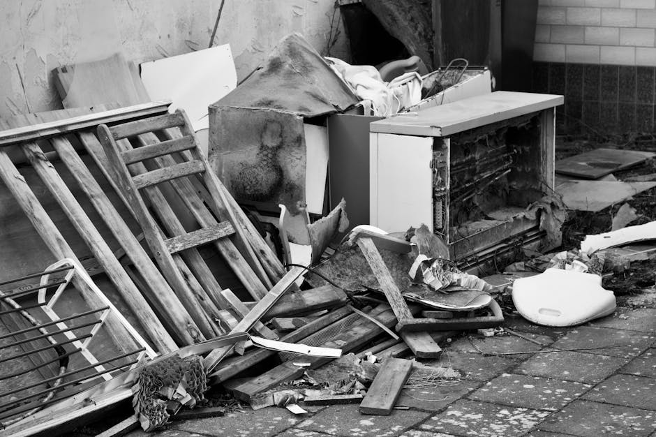A collection of discarded household waste and debris piled on a paved outdoor area, including a damaged wooden pallet leaning against a wall on the left side of the image, with weathered and splintered planks. Behind and partially covering the pallet, there is a rusty metal wire frame, possibly part of a shelving unit or container. In the center, a broken, partially dismantled white kitchen appliance or cabinet with exposed interior components and chipped edges is visible, topped with miscellaneous debris such as torn fabric or paper. To the right, a large, fractured white ceramic or plastic object, likely a bathroom fixture or container, rests on the ground. Surrounding the main objects are scattered wooden fragments, pieces of cardboard, and other trash, with some items leaning against a building wall constructed of brick and concrete blocks. The environment suggests a neglected or waste collection area, possibly intended for rubbish removal or disposal services, with natural light illuminating the clutter and broken objects.