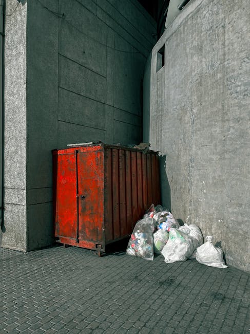 A large, rusty metal dumpster with a weathered red finish is positioned against the right side of a corner formed by a dark green wall and a light gray concrete wall. The dumpster's surface shows signs of wear, with patches of peeling paint and areas of discoloration. Several clear plastic bags filled with various types of rubbish, including plastic bottles, packaging, and mixed waste, are piled directly in front of the dumpster, some leaning against it. The scene is set outdoors on a paved surface made of dark, textured paving stones, with natural daylight illuminating the area. The environment appears to be a back alley or service area adjacent to modern buildings, indicating a private waste collection point rather than a public street. This setup reflects an alternative rubbish removal method typical of independent collection or on-site clearance services, as provided by companies like rubbishremovalwesthampstead.co.uk, serving areas such as NW6 in West Hampstead.