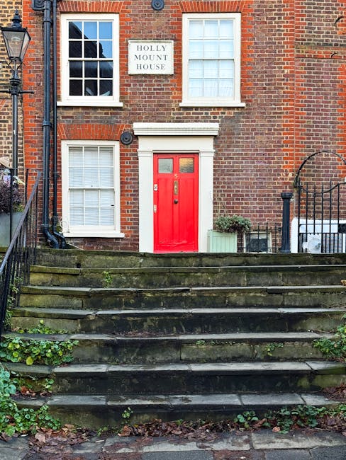 The image depicts the front entrance of a brick residential building with a red-painted wooden door situated centrally on a set of outdoor stone steps, which appear weathered with visible moss and leaves accumulated on the steps. To the left of the door, there is a large window with white framing and closed blinds, while on the right, a similar window with white trim is visible, both set into the red-brick facade. Above the door, there is a white decorative lintel, and to the right of the upper window, a sign reading 'HOLLY MOUNT HOUSE' is affixed to the brick wall. Surrounding the entrance area, a black metal railing runs along the sides of the stairs, with the right side slightly curved. A small green plant in a rectangular planter is positioned to the right of the door, next to a small shrub, suggesting some effort at decorative gardening. The environment indicates a quiet, well-maintained residential setting possibly in West Hampstead, with natural ambient light illuminating the scene and highlighting the brickwork texture and aged appearance of the steps, which could be part of an independent property or a private estate, relevant to the waste management context provided by rubbishremovalwesthampstead.co.uk.