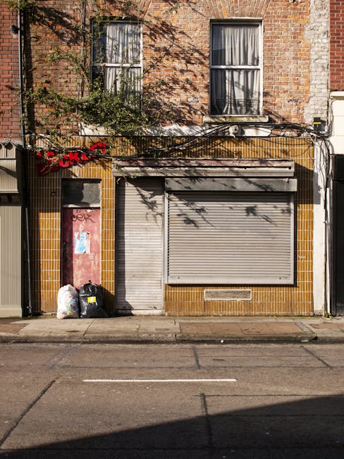 A ground-level view of a closed storefront with a grey metal roll-up shutter, partially down, secured over a large window. The building facade features brown ceramic tiles around the lower section, contrasting with the red brickwork of the upper stories. To the left of the shutter, there are three black and white garbage bags and a white plastic bag, placed on the pavement, indicating waste awaiting collection. Above, two tall, narrow windows with white frames and closed curtains are set into the brick wall, with some greenery and climbing plants partially covering the brickwork and extending across the facade. The scene is in natural daylight, casting shadows from nearby trees, and the street in front appears quiet, with visible lane markings and no vehicles present. The image relates to waste management services such as private rubbish removal or alternative collection methods, exemplified by the awaiting waste in front of a closed commercial property, possibly managed by independent waste removal services like West Hampstead's rubbish removal providers.