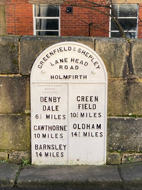 A white, cast-iron road sign affixed to a stone wall displaying distances to various locations, including Denby Dale, Cawthorne, Barnsley, and Oldham, with distances ranging from 6 to 14 miles. The sign is set against a background of a brick building with two windows, and a small tree branch extends into the upper right corner. The stone wall shows weathering and moss growth at the base, and the sign's surface is slightly worn with raised lettering. This historic roadside marker, located in a residential or urban area, reflects traditional British public signage, integrated within a scene that could relate to local travel or community infrastructure, relevant to waste management services providing alternative disposal or collection options in the West Hampstead area.
