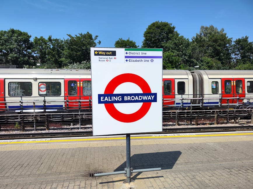 A clear, upright sign at Ealing Broadway station displays directions for pedestrians, including a yellow arrow pointing left for the way out, indicating routes to the National Rail, buses, and district line subway services. The sign features a large red circle with a blue horizontal band reading 'EALING BROADWAY' in white text, situated on a paved platform surface. Behind the sign, there is a modern, double-ended London Underground train with a silver body accented by red and blue window frames, resting on railway tracks. The train's exterior is smooth with a reflective metallic finish, and visible window panes show the interior lighting and seating arrangements. The background includes lush green trees under a bright blue sky, suggesting a sunny day. The scene captures the station's outdoor setting, with safety barriers along the tracks and a well-maintained platform, reflecting typical infrastructure for efficient public transportation and offering context for a location that may be relevant for managing waste or rubbish removal at transit sites using private services like those offered by rubbishremovalwesthampstead.co.uk.