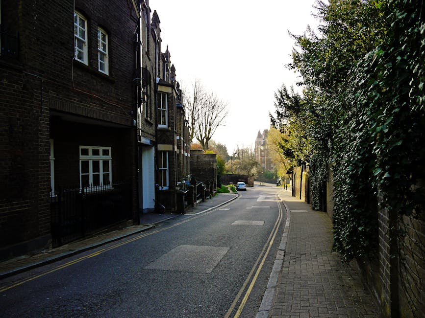 A quiet residential street scene featuring a narrow tarmac road with double yellow lines along the curb on the left side, and a paved sidewalk on the right, bordered by dense greenery and a brick wall. On the left side, there are dark brick terraced houses with white window frames and small front gardens, some enclosed by black metal fences. The street is illuminated by soft daylight with shadows cast by trees and buildings, and a single white car parked further along the road. The environment appears calm and typical of urban West Hampstead, with no visible rubbish or waste materials present. The scene subtly suggests the context of local, independent rubbish removal services like those provided by [COMPANY_NAME], highlighting the importance of efficient waste handling in maintaining tidy residential areas.